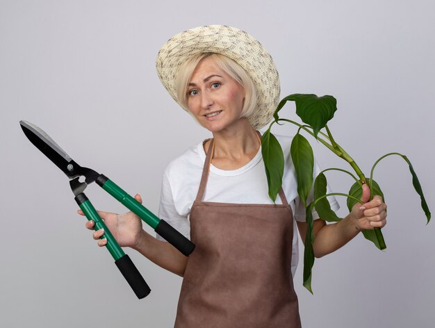 Photo gratuite femme souriante de jardinier blonde d'âge moyen en uniforme portant un chapeau tenant une plante et des cisailles à haie isolées sur un mur blanc
