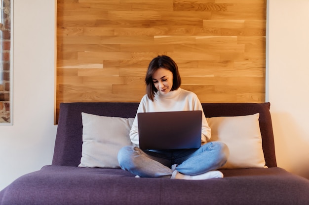 Femme souriante en jeans et t-shirt blanc travaille sur un ordinateur portable assis sur un lit sombre devant un mur en bois à la maison