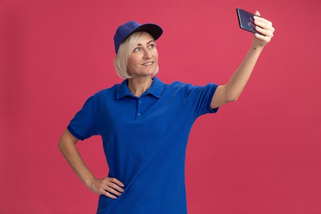 Femme souriante de livraison blonde d'âge moyen en uniforme bleu et casquette gardant la main sur la taille prenant un selfie isolé sur un mur rose