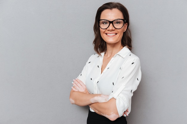 Femme souriante à lunettes et vêtements d'affaires posant avec les bras croisés et regardant la caméra sur fond gris