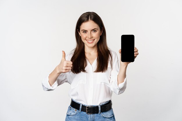 Femme souriante montrant les pouces vers le haut, recommandant l'application, l'écran du téléphone portable, l'interface vide sur le smartphone, debout sur fond blanc.