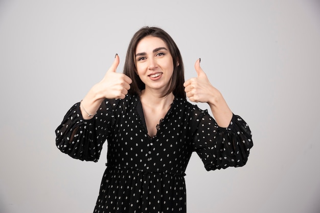 Femme souriante montrant les pouces vers le haut et regardant la caméra sur fond gris.