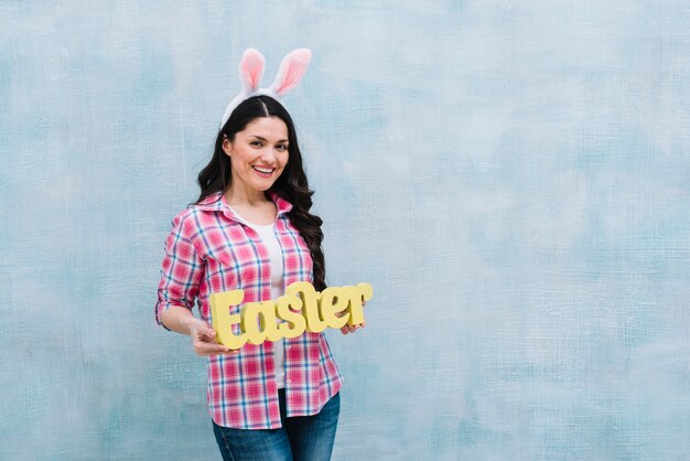 Femme souriante avec oreille de lapin montrant le mot de Pâques sur fond texturé bleu