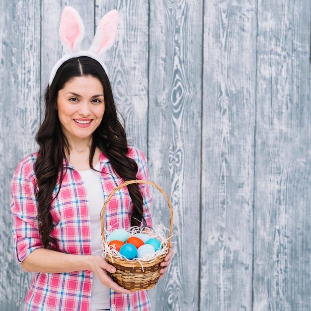 Femme souriante avec des oreilles de lapin sur la tête montrant le panier d&#39;oeufs de Pâques sur un fond en bois