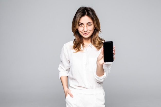 Femme souriante pointe sur smartphone debout sur un mur blanc.