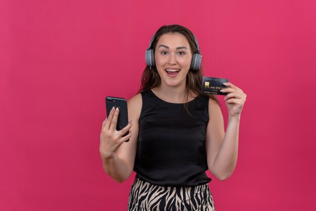 Femme souriante portant maillot noir portant des écouteurs tenant le téléphone et la carte bancaire sur le mur rose