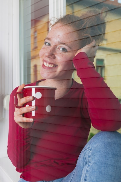 Femme souriante regardant à travers la fenêtre et dégustant un café