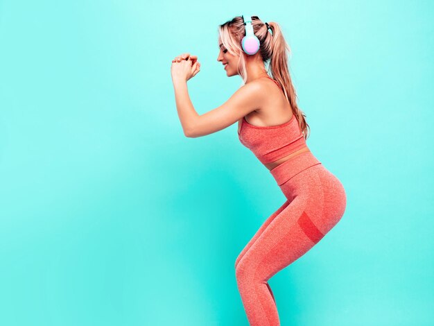 Femme souriante de remise en forme dans des vêtements de sport roses Jeune beau modèle avec un corps parfait Femme posant près du mur bleu en studio Joyeux et heureux Écouter de la musique dans des écouteurs sans fil Faire des squats
