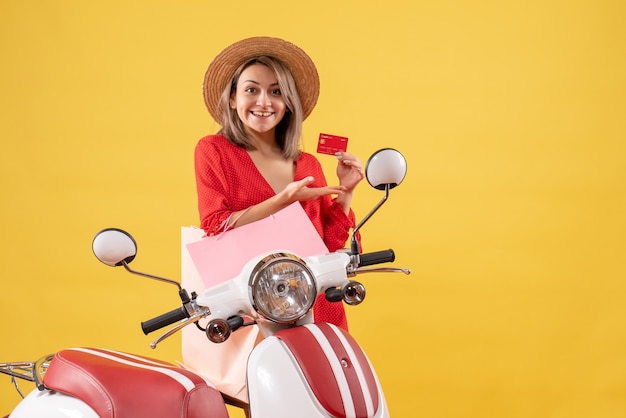 femme souriante en robe rouge sur un cyclomoteur tenant des sacs à provisions et une carte