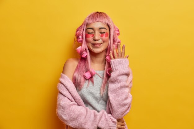 Femme souriante satisfaite aux cheveux roses, applique des rouleaux et des coussinets de beauté, vêtue d'un pull rose, profite de temps libre pour se dépenser seule