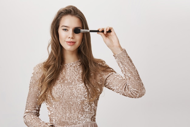 Femme souriante séduisante en robe de soirée, couvrir les yeux avec un pinceau de maquillage