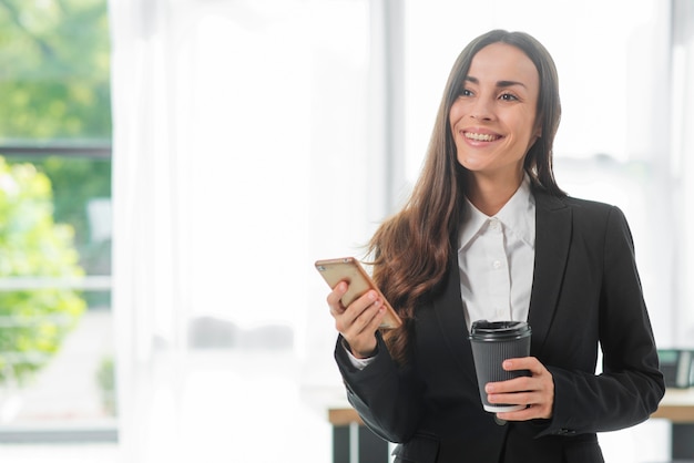 Femme souriante tenant un smartphone et une tasse de café jetable