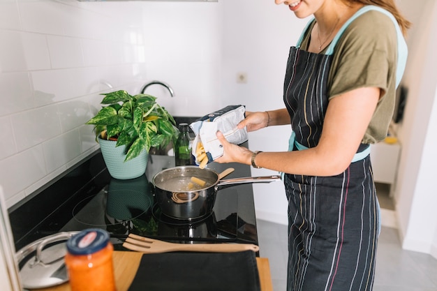 Photo gratuite femme souriante, verser des pâtes rigatoni dans la casserole sur la cuisinière électrique