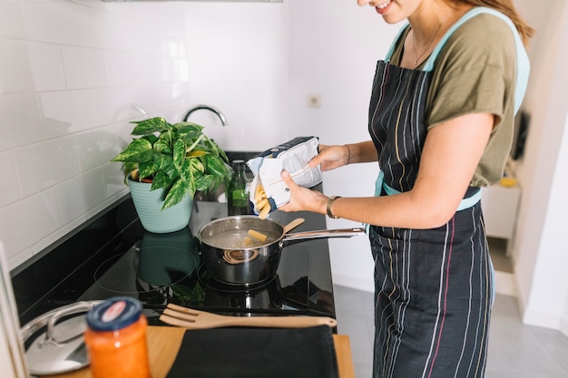 Femme souriante, verser des pâtes rigatoni dans la casserole sur la cuisinière électrique