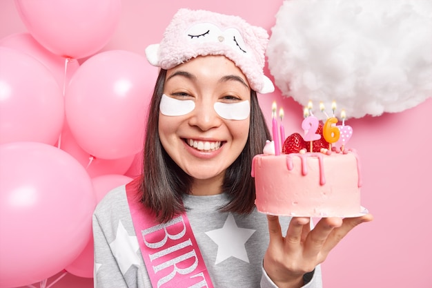 La femme sourit largement détient un délicieux gâteau d'anniversaire aime la célébration à la maison vêtue de vêtements domestiques décontractés pose sur rose