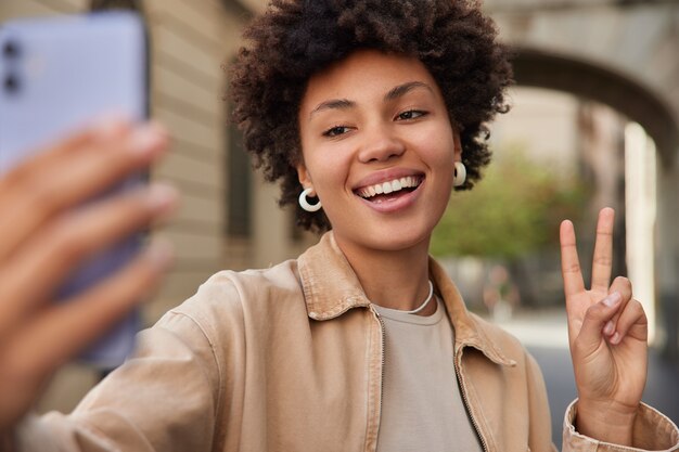 une femme sourit largement fait un portrait de sefie montre un signe de paix sourit largement se promène en ville vêtue de vêtements élégants profite de ses loisirs en milieu urbain