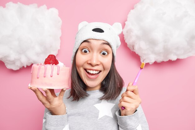 une femme sourit à pleines dents vêtue d'un pyjama tient un délicieux gâteau et une brosse à dents a des caries parce qu'elle mange trop de bonbons isolés sur rose