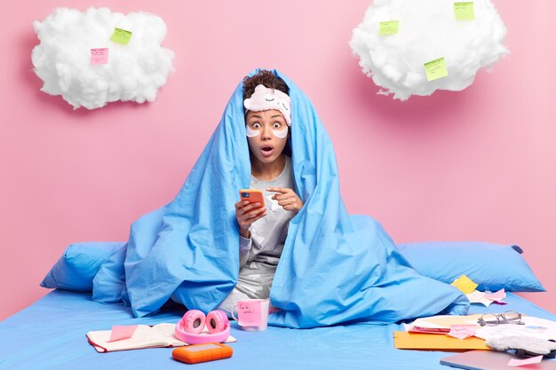 une femme sous une couverture pointe sur un smartphone découvre des nouvelles choquantes passe du temps libre à la maison porte un masque de sommeil et un pyjama fait des devoirs à distance