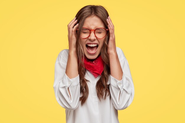 Une femme sous pression et fatiguée tient les mains sur la tête, a mal à la tête ou migraine, garde la bouche largement ouverte tout en s'exclamant quelque chose