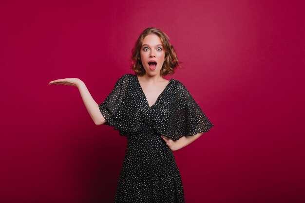 Femme spectaculaire en écharpe tricotée à la mode couvrant les yeux avec les mains. Photo intérieure d'une femme séduisante en riant en chapeau et pull drôle posant devant un mur violet.