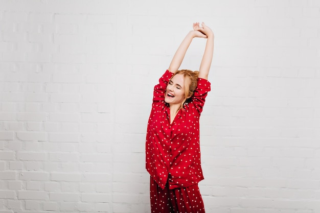 Femme spectaculaire en vêtements de nuit à la mode qui s'étire avec le sourire. Fille enthousiaste appréciant bonjour et posant sur un mur blanc.