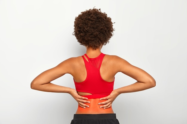 Photo gratuite une femme sportive active s'est blessée au dos après avoir fait de l'exercice ou de la course, ressent une sensation de douleur dans le bas du dos, a les cheveux bouclés, est vêtue d'un haut et d'un pantalon rouges