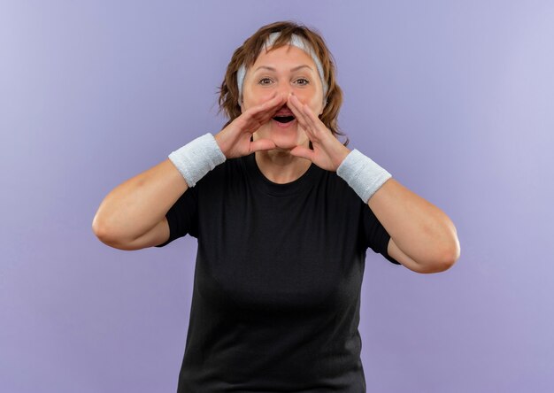 Femme sportive d'âge moyen en t-shirt noir avec bandeau en criant ou en appelant quelqu'un avec les mains près de la bouche debout sur le mur bleu