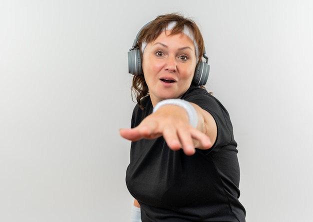 Femme sportive d'âge moyen en t-shirt noir avec bandeau et écouteurs à la surprise en pointant avec bras debout sur mur blanc