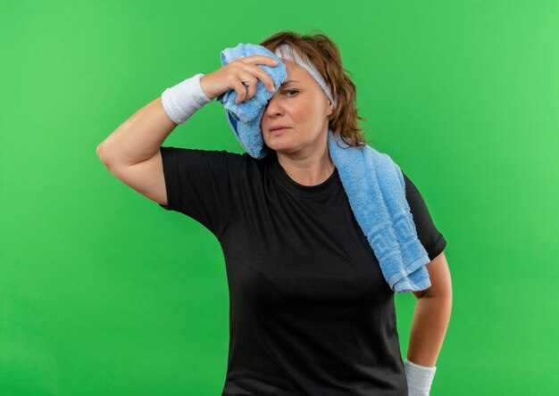 Femme sportive d'âge moyen en t-shirt noir avec bandeau et serviette sur son cou à la fatigue et épuisée après l'entraînement debout sur le mur vert