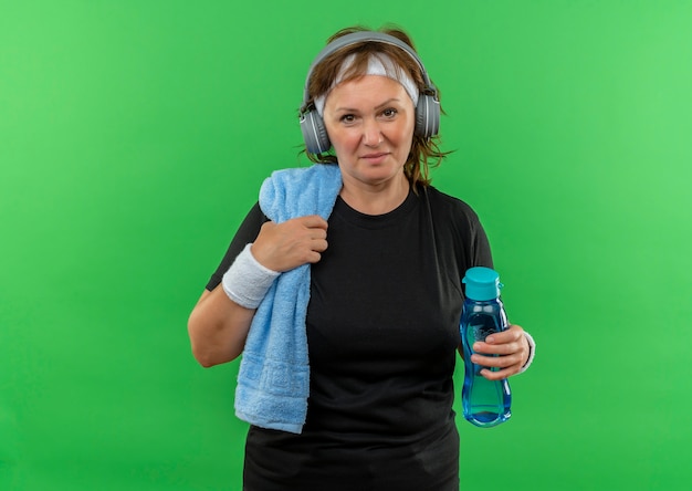 Photo gratuite femme sportive d'âge moyen en t-shirt noir avec bandeau et serviette sur son cou tenant une bouteille d'eau fatiguée et heureuse après le travail debout sur un mur vert