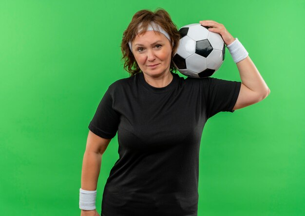 Femme sportive d'âge moyen en t-shirt noir avec bandeau tenant un ballon de football avec une expression sérieuse et confiante debout sur un mur vert