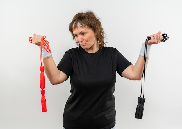 Femme sportive d'âge moyen en t-shirt noir avec bandeau tenant deux cordes à sauter à la confusion ayant des doutes debout sur un mur blanc