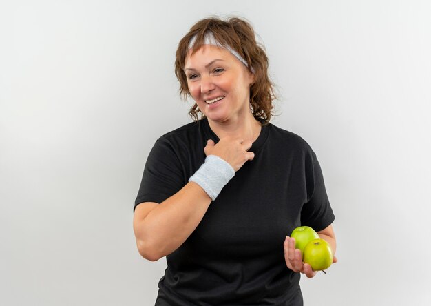 Femme sportive d'âge moyen en t-shirt noir avec bandeau tenant deux pommes vertes souriant joyeusement debout sur un mur blanc