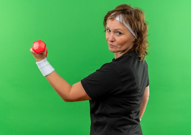 Femme sportive d'âge moyen en t-shirt noir avec bandeau travaillant avec haltère à la confiance debout sur mur vert