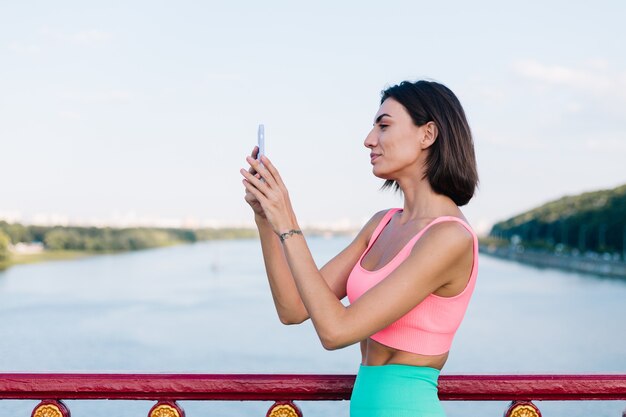 Femme sportive dans des vêtements de sport adaptés au coucher du soleil sur un pont moderne avec vue sur la rivière sourire positif heureux avec téléphone portable