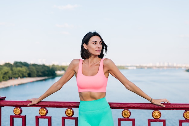 Femme sportive dans des vêtements de sport adaptés au coucher du soleil sur un pont moderne avec vue sur la rivière sourire positif heureux