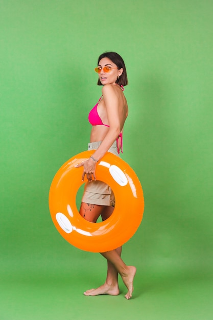 Femme sportive en forme d'été en bikini rose et anneau gonflable orange vif rond et lunettes de soleil sur vert, joyeux joyeux excité joyeux positif