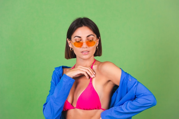 Femme sportive en forme d'été en bikini rose, chemise bleue et lunettes de soleil orange sur vert, joyeux joyeux joyeux positif