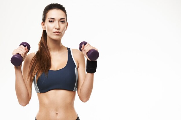 Femme sportive avec des haltères en tenue de sport posant sur blanc.