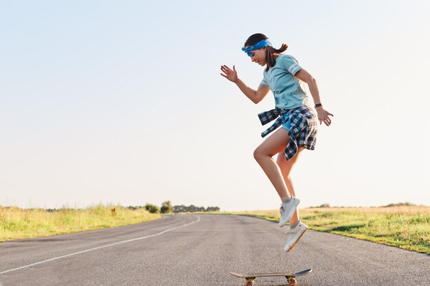 Femme sportive portant un t-shirt et un short faisant des tours sur une planche à roulettes dans la rue sur une route goudronnée, sautant en l'air, profitant de la planche à roulettes seule au coucher du soleil en été.