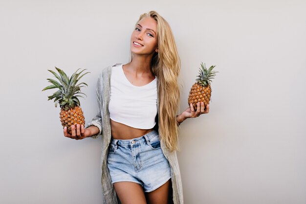 Femme sportive de rêve à la peau bronzée posant sur fond clair. Plan intérieur d'une femme blonde incroyable avec des ananas.