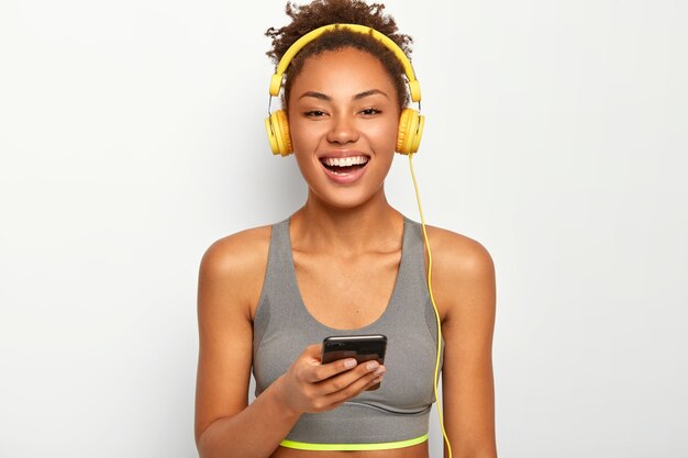 Femme sportive en vêtements de sport, sourit largement, utilise un téléphone portable moderne, écoute de la musique via des écouteurs, pose sur fond blanc.