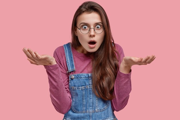 Une femme stupéfaite incertaine a horrifié l'expression désemparée, écarte les mains, regarde avec choc, porte des lunettes