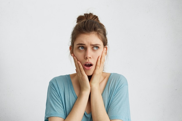 Femme stupéfaite avec noeud de cheveux portant un chemisier bleu décontracté tenant les mains sur les joues ouvrant sa bouche avec surprise en levant