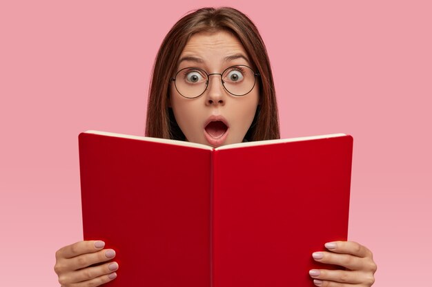 Une femme stupéfaite regarde un livre avec des yeux bouchés et une bouche ouverte, porte des lunettes transparentes, surprise par le contenu étonnant de l'histoire, ne peut pas croire à une fin choquante, des modèles sur un mur rose