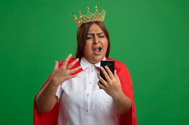 Femme de super-héros d'âge moyen en colère portant couronne tenant et regardant le téléphone isolé sur vert