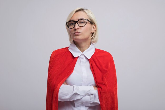 Femme de super-héros blonde d'âge moyen confiante en cape rouge portant des lunettes debout avec une posture fermée isolée sur un mur blanc avec espace de copie