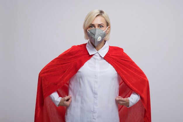 Femme de super-héros blonde d'âge moyen impressionnée en cape rouge portant un masque de protection debout comme un surhomme isolé sur un mur blanc