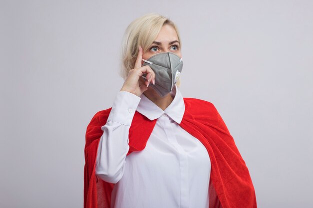 Femme de super-héros blonde d'âge moyen impressionnée en cape rouge portant un masque de protection regardant de côté faisant un geste de réflexion isolé sur un mur blanc avec espace de copie