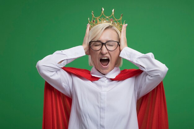 Femme de super-héros blonde d'âge moyen stressée en cape rouge portant des lunettes et une couronne gardant les mains sur la tête en criant les yeux fermés isolés sur un mur vert